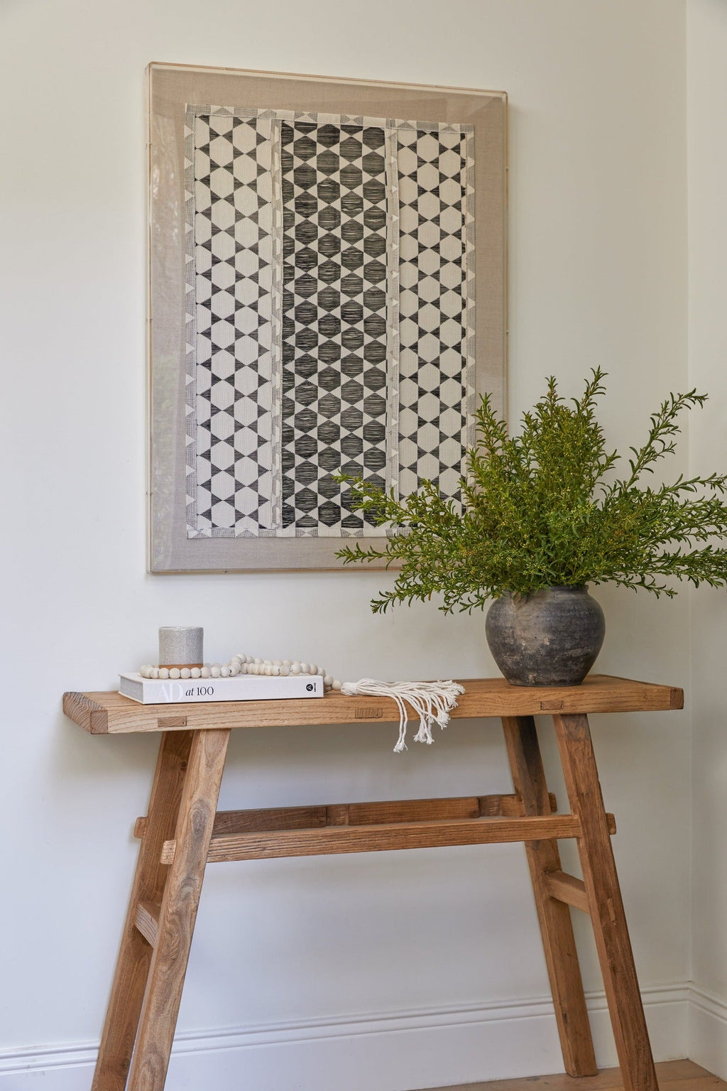 Antique clay pot with greenery on wooden table under patterned wall art in minimalist room