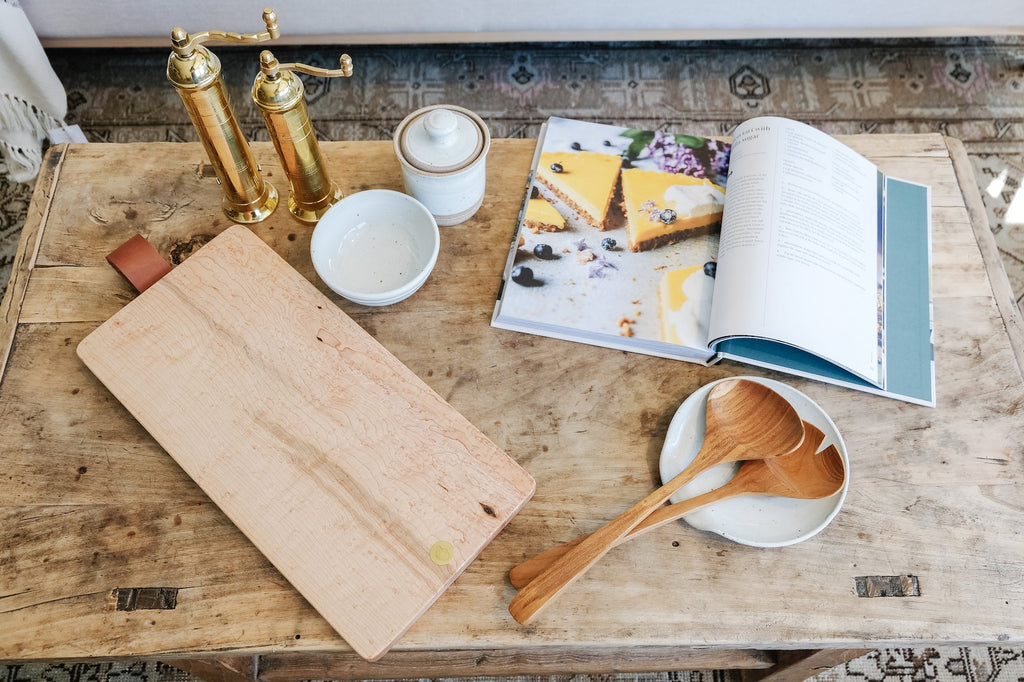 Hand-carved teak salad servers on a rustic wooden table with cutting board, bowls, and open cookbook