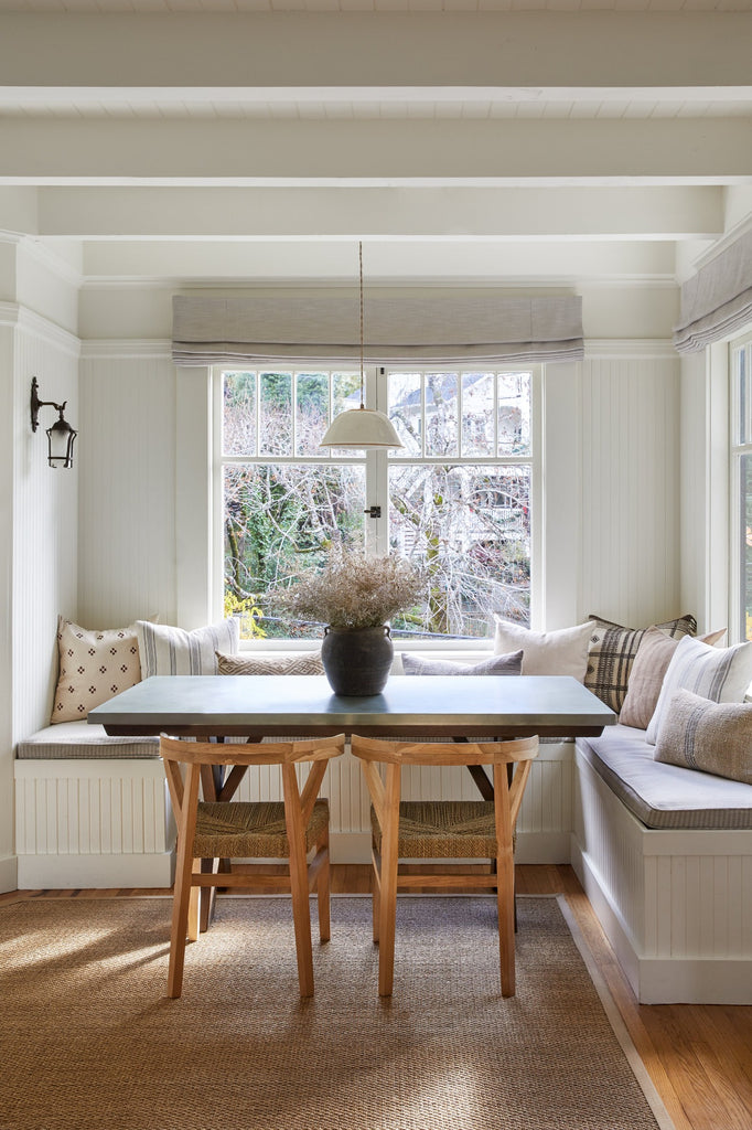 Hand thrown ceramic light pendant in a breakfast nook with brown checkered pillow, Buhjodi pillow, antique clay pot, and woven wishbone dining chairs. -Saffron + Poe 