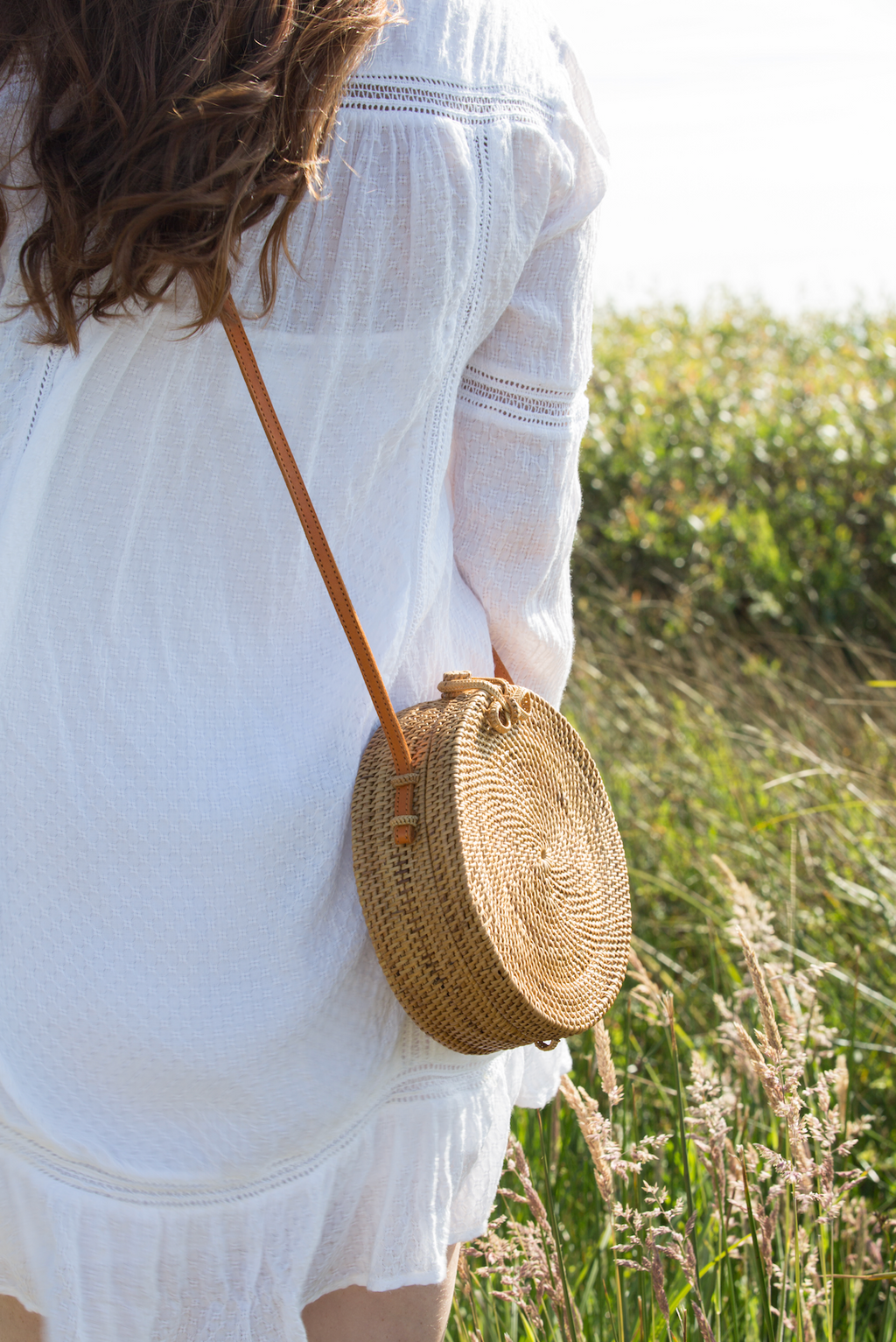 Woman wearing a white dress carrying a round woven Tenganan Basket Crossbody bag with a leather strap outdoors.