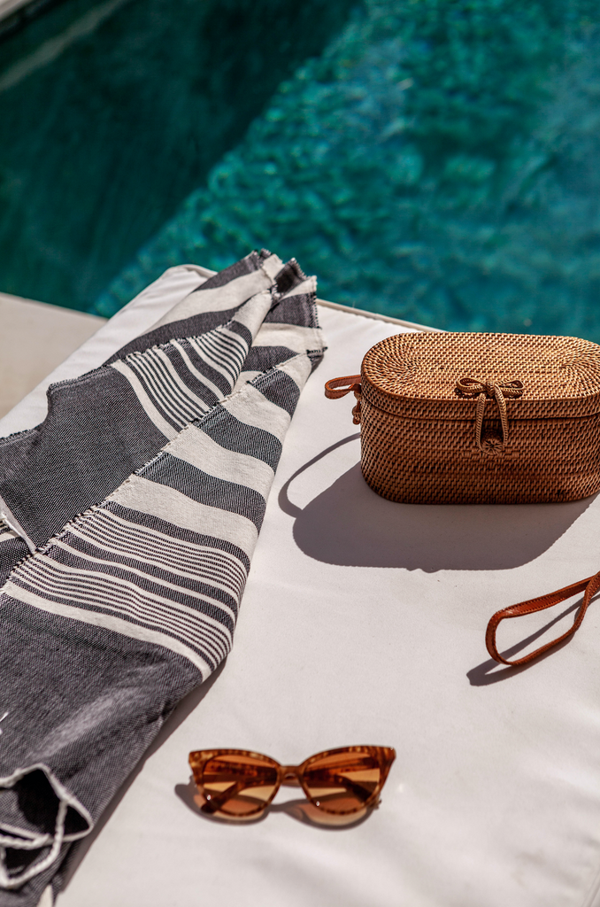 Styled laydown photo next to the pool with our Tenganan Basket Crossbody Bag - oval, small and chic. Handmade in Tenganan, Bali using natural Ata reed. - Saffron and Poe
