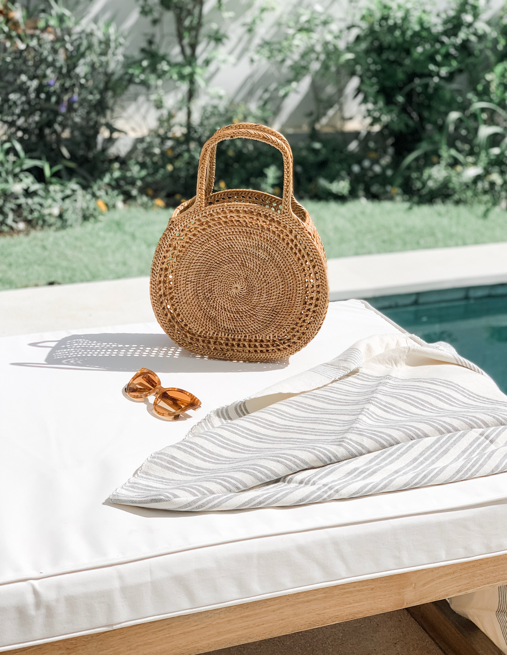 Tenganan Basket Handbag round woven Ata reed bag on white lounge chair with sunglasses and striped cloth near pool outdoors