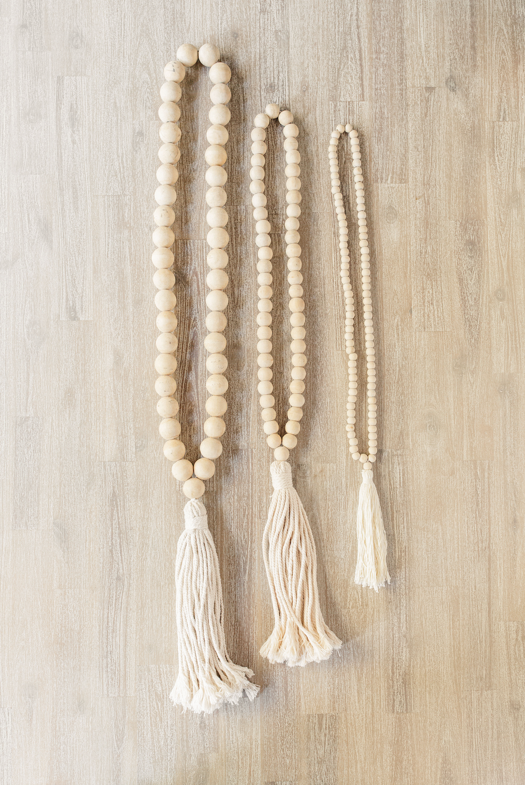 Three sizes of tasseled wooden Bali beads with cream cotton tassels displayed on a wooden surface