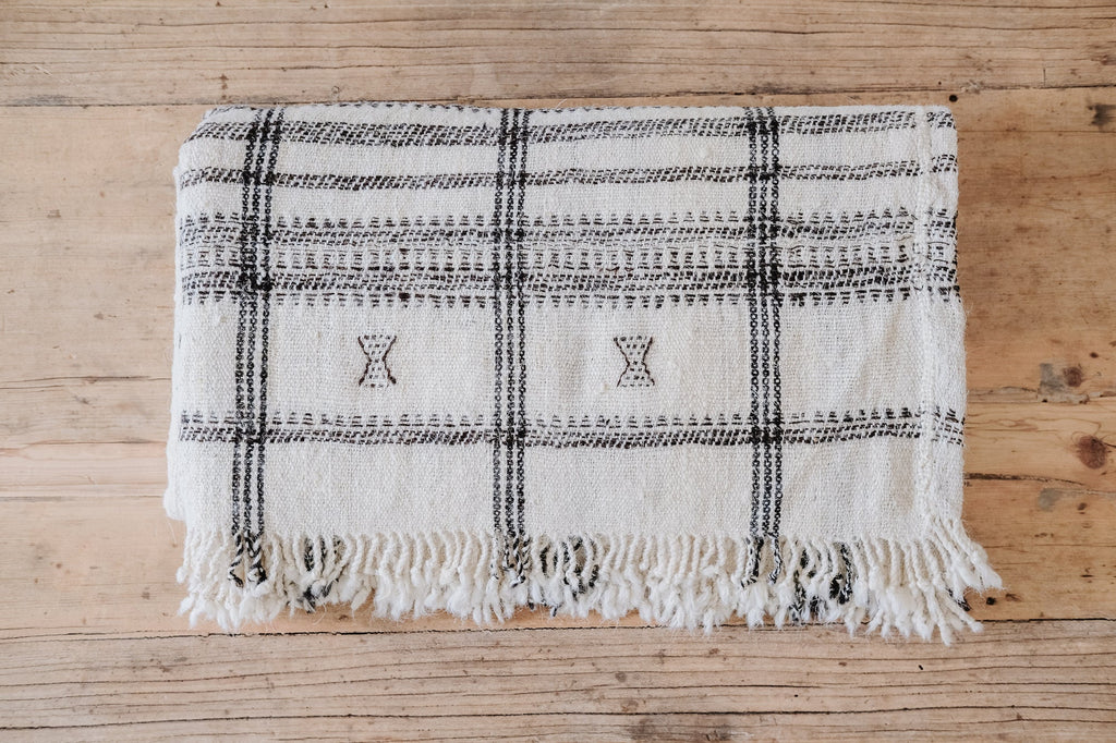 Handwoven Bhujodi Blanket in natural tones with black woven pastoral motifs and fringed edges on wooden surface