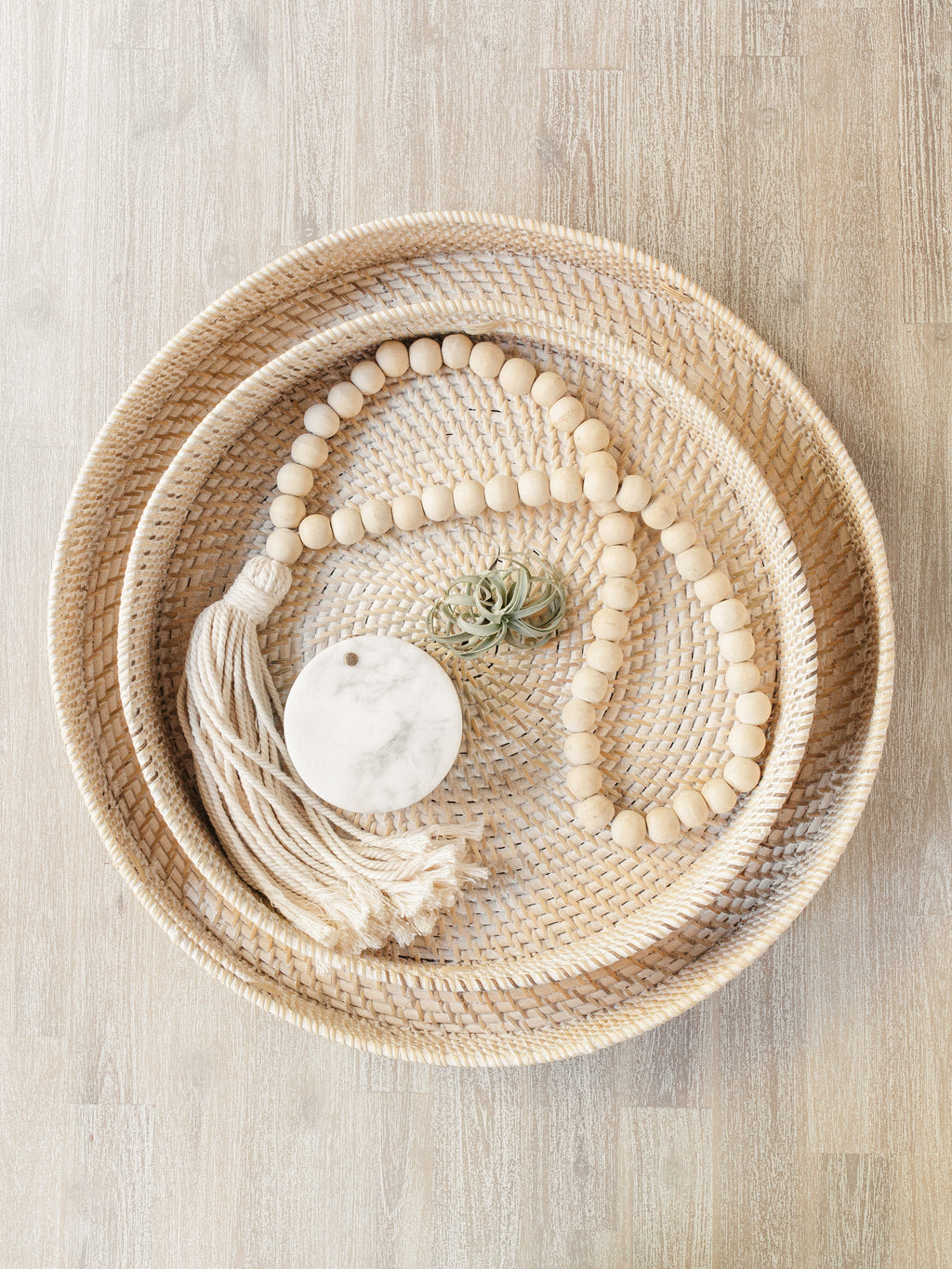 Tasseled wooden Bali beads with cream cotton tassel displayed on woven trays with air plant and marble coaster on wooden surface