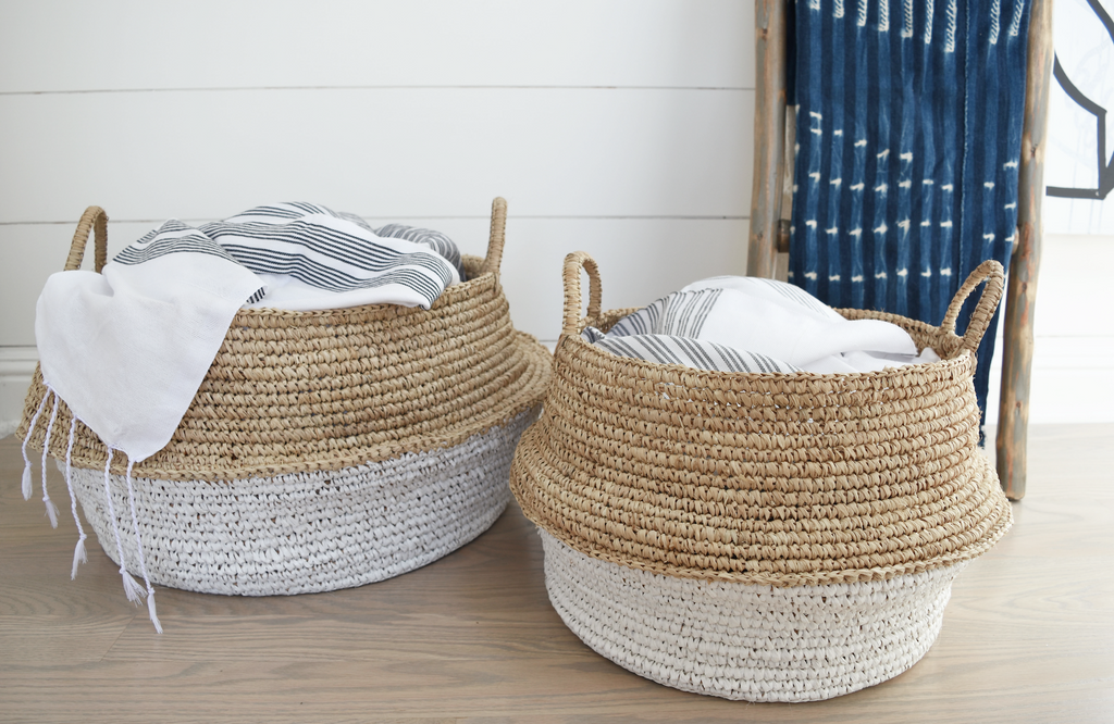 Two-toned collapsible woven hyacinth baskets with handles, filled with white and striped textiles, displayed on a wooden floor.