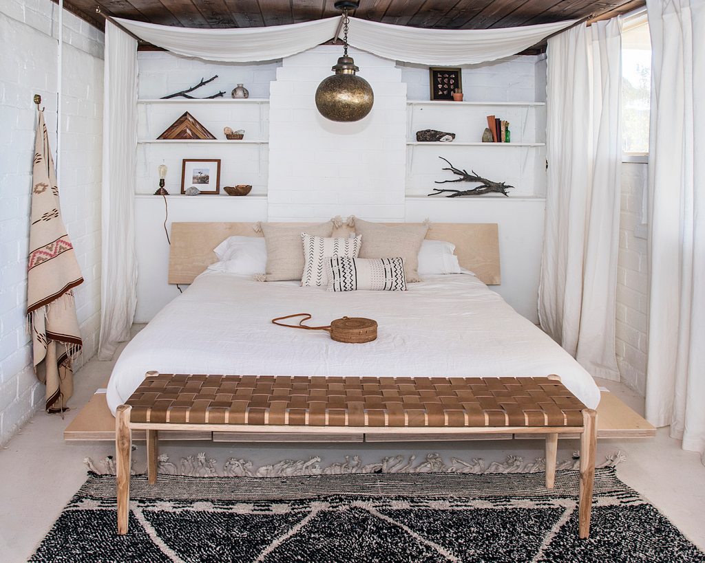 Woven leather bench with teak frame and saddle leather straps at the foot of a white bed in a rustic bedroom