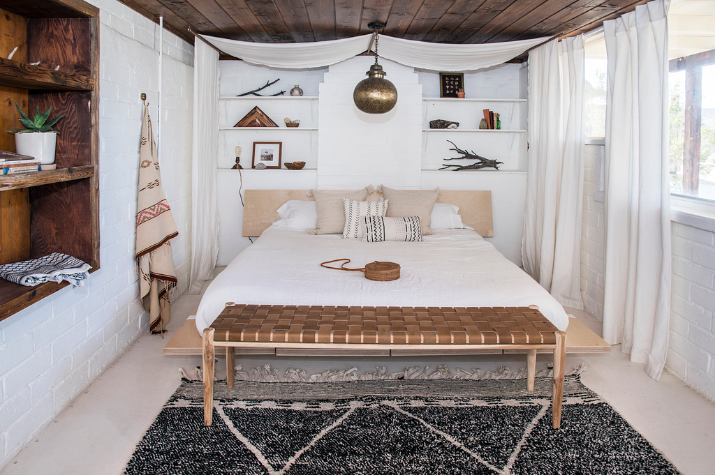 Woven leather bench with teak frame and saddle leather straps at the foot of a white bed in a rustic bedroom